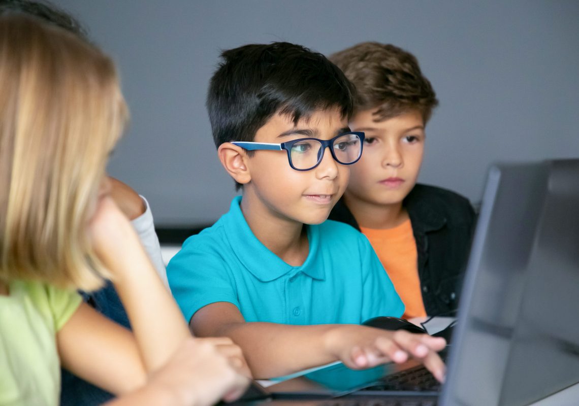 Asian boy typing on laptop keyboard and classmates sitting at table, watching him and doing task together. Pupils using computers during study. Childhood, communication and digital education concept